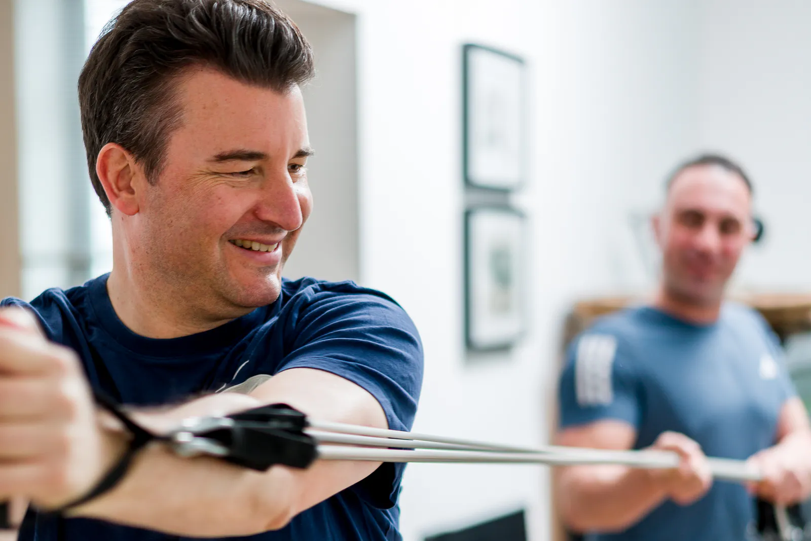 Ein Klient trainiert lächelnd mit einem Expander in seiner Wohnung, im Hintergrund leitet Personal Trainer Nico Fichtler an